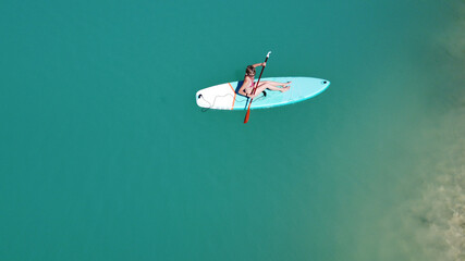 A girl in a dress floats on a glanders board on a pond with bright turquoise water. Warm summer day for travel. Top view from a quadcopter. Aerial photography