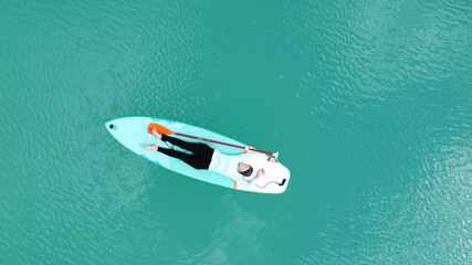 A girl in a dress floats on a glanders board on a pond with bright turquoise water. Warm summer day for travel. Top view from a quadcopter. Aerial photography