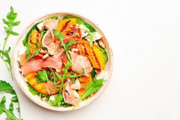 Salad bowl with grilled peach, prosciutto, goat cheese, walnuts and arugula on white background, top view, copy space