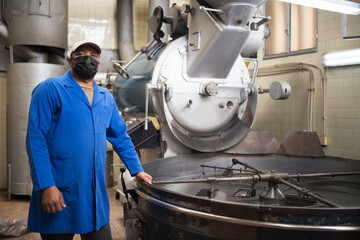 Mid adult workman standing next to coffee roasting machine. Interior of coffee production workshop with set up roasting equipment. Person in uniform and mask. Production, food concept