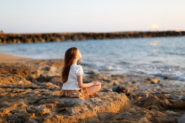 Cute caucasian child girl with long hair meditates by the sea, silence and calmness, warm toning