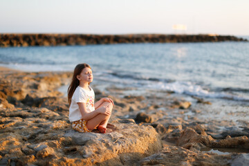 Fototapeta na wymiar Cute caucasian child girl with long hair meditates by the sea, silence and calmness, warm toning