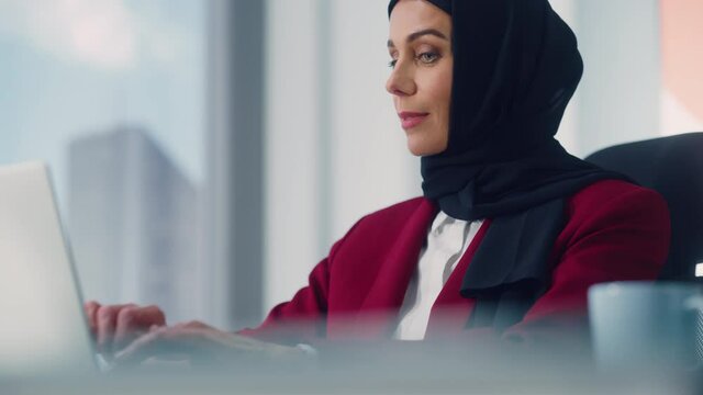 Close-up Portrait of Gorgeous Muslim Businesswoman Wearing Hijab Sitting at Her Desk Working on Laptop Computer in Office. Successful Corporate CEO Plan Investment Strategy for e-Commerce Startup