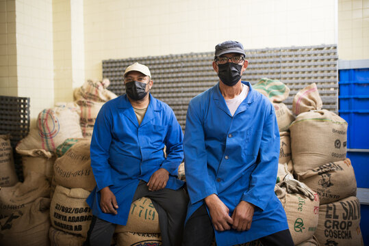 Portrait of two coffee roasters at work. Men sitting at jute sacks. Men in face mask looking at camera. Job, factory, maintenance concept