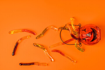 A pumpkin-shaped pot is filled with marmalade worms on an orange background. The funny concept of Halloween.
