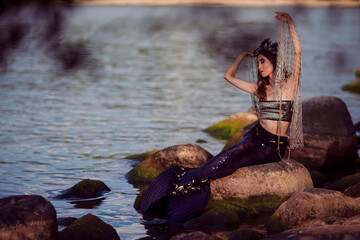 Asian Mermaid Embracing With Net At Sea Shore on Rocks While Wearing Seashell Decorated Crown and Black Shiny Tail On Sexy Body Covered With Strasses As Mistress Queen of Sea.