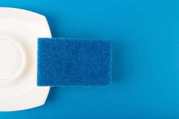 Dishwashing minimal creative concept. Close up of white shiny clean plate and blue cleaning sponge against blue background with copy space. 