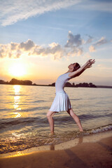 Fototapeta na wymiar Sensual Japanese Ballet Dancer in White Dress And Silver Crown Posing In Ocean Waves While Showing Ballet Pas With Lifted Hands in Dance Pose On Picturesque Sunset At Sea