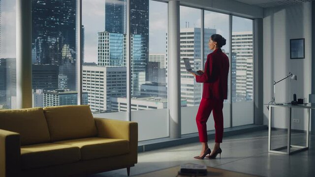 Successful Caucasian Businesswoman Using Laptop While Standing in Office Looking out of Window on Big City. Confident Female Corporate Top Manager of IT Company Plans Investment Strategy