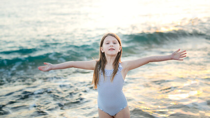 Fototapeta na wymiar Cute caucasian child girl in a striped swimsuit with long hair with outstretched arms hugs the sea on an empty beach at a resort, evening and sunset
