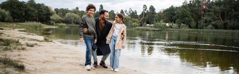 Smiling arabian man hugging kids near lake in park, banner