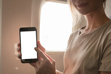 Mockup image of a woman holding and looking at smart phone with blank white screen next to an airplane window with clouds and sky background