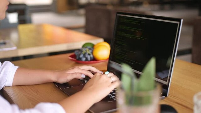 Back view over the shoulder shot of developer programmer female with laptop. Program code and script data on the screen. Young freelancer woman working on project in cafe.