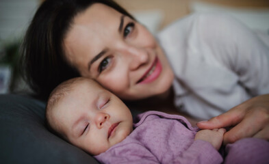 Front view of happy young mother with her newborn baby girl lying on sofa indoors at home, looking at camera.