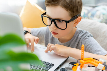 Clever young boy in glasses working on a laptop for his new project, programs a robotic vehicle, codes an electronic toy at home. STEM education and programming lesson concept.