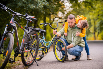 Dad and daughter inspect wheel of children's teenage bicycle on the autumn path of the park.