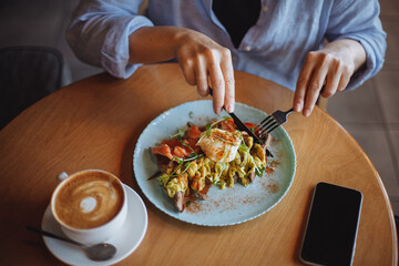 middle-aged woman in a blue shirt eats scrambled eggs and cappuccino for breakfast in a cafe