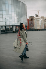 Portrait of smiling young brunette girl in multicolored scarf and olive trench coat in autumn city