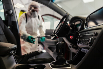 Naklejka premium COVID 19 situation warning. A man in a protective white uniform and a sprayer prevents the spread of the infection inside of a car, showroom. Stay responsible, wear the mask, coronavirus, virus