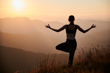 Back view of young woman performing yoga pose on grassy hill with orange sky on background. Fit...