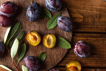 Ripe plums on a wooden cutting board, among green leaves and twigs. One plum is cut in half and the pulp and bone are visible.
