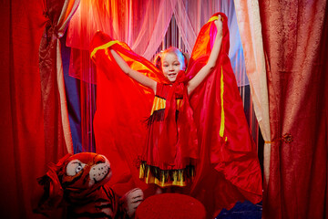 Small girl during a stylized theatrical circus photo shoot in a beautiful red location. Young model posing on stage with curtain