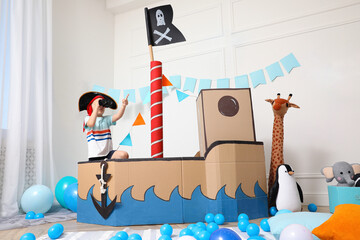 Little boy playing with binoculars in pirate cardboard ship at home. Child's room interior
