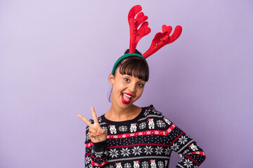 Young mixed race woman with reindeer hat celebrating Christmas isolated on purple background  joyful and carefree showing a peace symbol with fingers.
