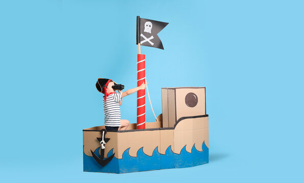 Little boy playing with binoculars in pirate cardboard ship on turquoise background
