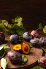 Ripe plums on a wooden cutting board, among green leaves and twigs. One plum is cut in half and the pulp and bone are visible.