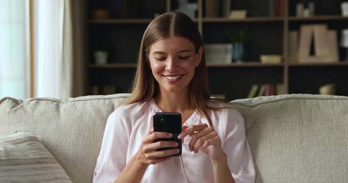 Pretty teenage female spend time at home communicate online look at mobile phone screen text message in chat to boyfriend read answer with smile. Happy young woman contact friend at web messenger app
