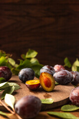 Fototapeta premium Ripe plums on a wooden cutting board, among green leaves and twigs. One plum is cut in half and the pulp and bone are visible.