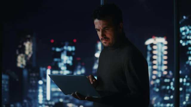 Big City Modern Office at Night: Successful Businessman Standing and Using Laptop. Handsome Male Digital Entrepreneur Thinking of Investment Strategy for e-Commerce Project. Medium Shot