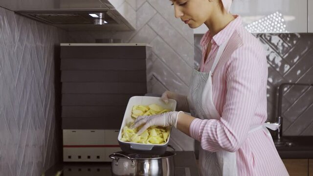 Attractive caucasian woman in apron and gloves shifting slices of apples from baking dish into pan. Young blonde using organic ripe fruits for bakery on modern kitchen.