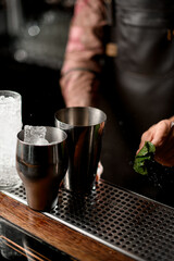 view of steel bar surface with steel shaker glasses with ice cubes and male hand with mint
