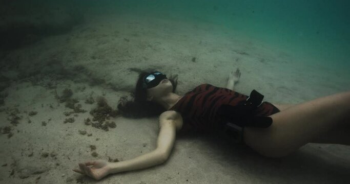 Crazy freediver woman lying underwater bottom, filmed on cinema camera. Brutal girl in black suits against background of mountains and ocean. Beautiful brunette touching her shoulder. Concept of