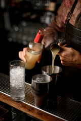 hand of bartender holds bottle and other hand pours drink from jigger into shaker glass