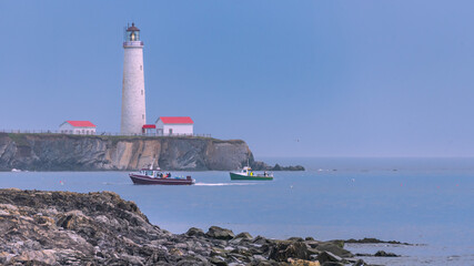 Canadian East Coast lobster fishing boats with Cap-des-Rosiers lighthouse in the background. - obrazy, fototapety, plakaty