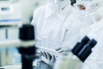 Middle shot of two health care scientists, wearing protective white gear, working in the laboratory. Researchers looking at a tablet, analyzing and evaluating data on the screen.