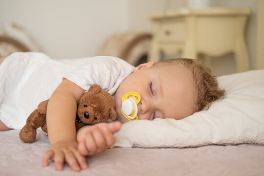 cute baby boy with nipple sleeping on bed at home. child hugging teddy bear. 