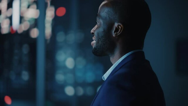 Night Office: Stylish Close-up Portrait of Powerful Black Businessman Wearing Suit Standing, Looking out of the Window on a Big City. Ambitious African CEO Thinking of e-Commerce Investment