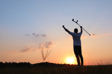 Man raising elbow crutch up to sky outdoors at sunset, back view and space for text. Healing miracle
