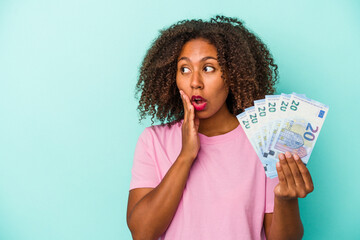 Young african american woman holding euro banknotes isolated on blue background is saying a secret hot braking news and looking aside