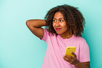 Young african american woman holding a mobile phone isolated on blue background touching back of head, thinking and making a choice.