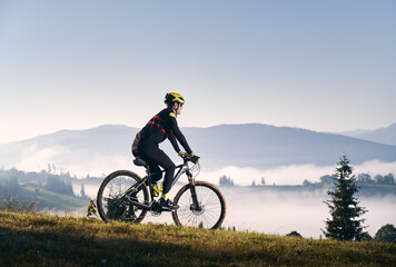 Naklejka na ściany i meble Side view of cyclist in cycling suit riding bicycle on grassy hill and looking at beautiful misty mountains. Male bicyclist in safety helmet enjoying the view of majestic mountains during bicycle ride