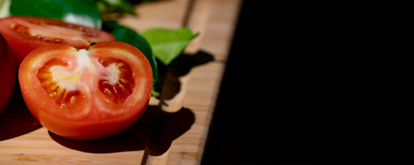Tomato and sliced half of tomato beside it, on wood plate in studio light with dark theme.