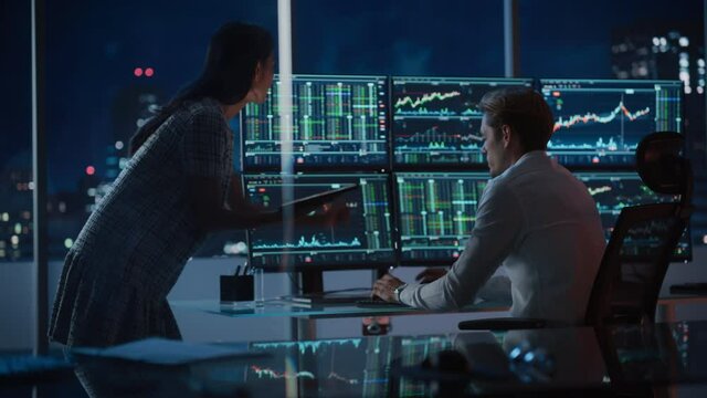 Financial Analyst Talking to Investment Banker in the Evening at Work. Chatting About Real-Time Stock Chart Data on Multi-Monitor Workstation. Businesspeople Have a Meeting in Broker Agency Office.