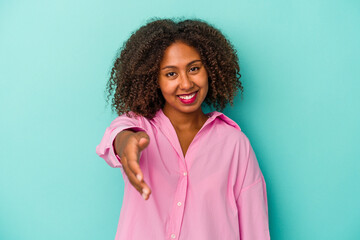 Fototapeta premium Young african american woman with curly hair isolated on blue background stretching hand at camera in greeting gesture.