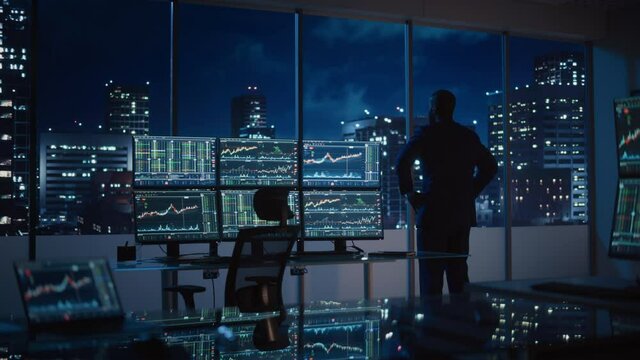 Successful Businessman Looking Out of the Window on Late Evening. Modern Hedge Fund Office with Computer with Multi-Monitor Workstation with Real-Time Stocks, Commodities and Exchange Market Charts.
