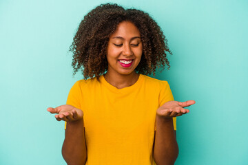 Young african american woman with curly hair isolated on blue background holding something with palms, offering to camera.